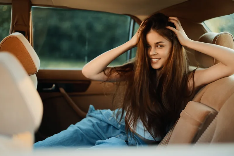 Young woman with long hair smiling in a car, wearing casual clothing, evoking a carefree and relaxed mood, with a natural green background.