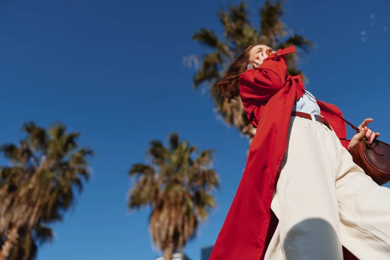A woman in a bold red coat walks against a bright blue sky with tall palm trees in the background.