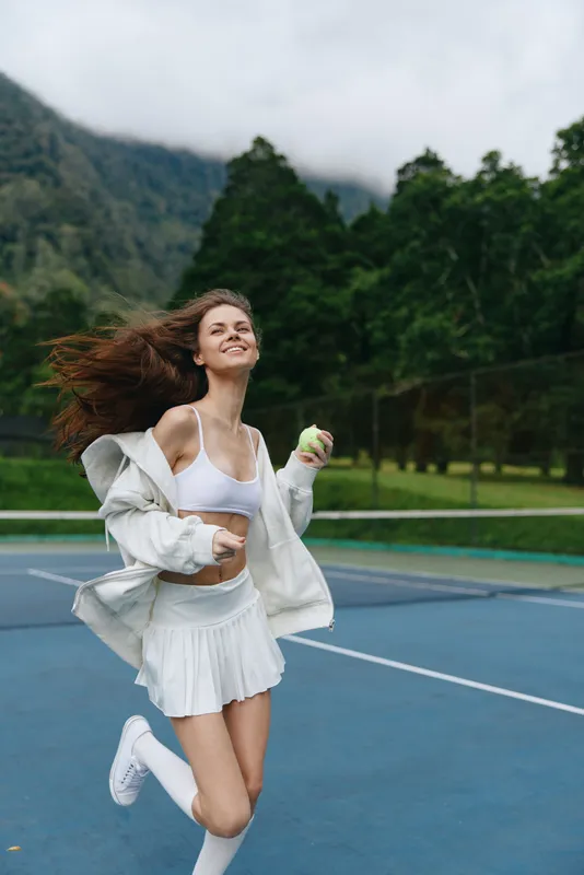 Happy young woman playing tennis outdoors, wearing a sporty outfit with a white jacket and skirt, showcasing joy and activity amidst a natural background