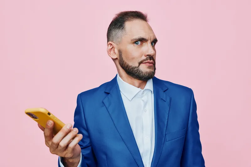A professional man in a blue suit stands against a solid pink background, displaying thoughtful intensity and calm focus, while holding a smartphone at chest level for a modern, corporate vibe.