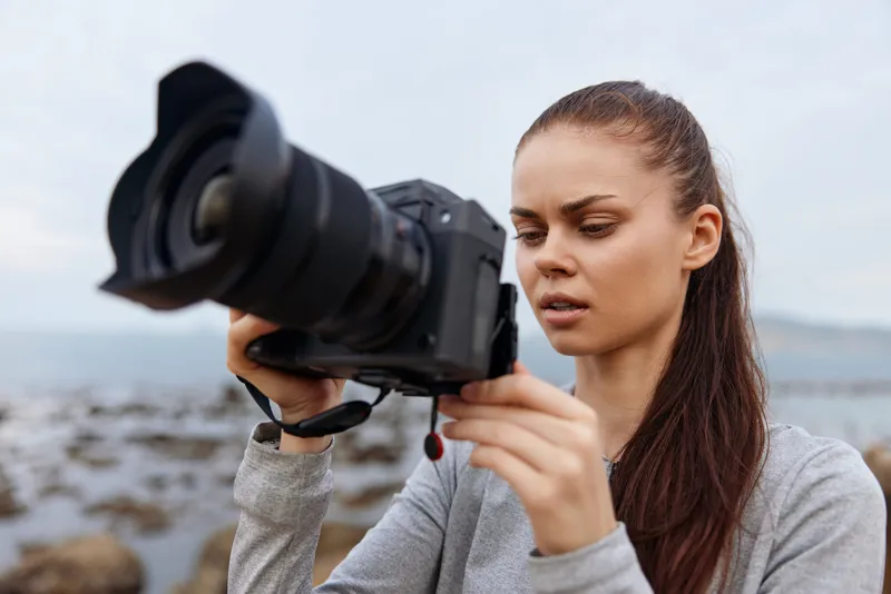 Young woman portrait taking photos by the sea, wearing casual grey outfit, showing concentration and passion for photography amid a serene natural backdrop