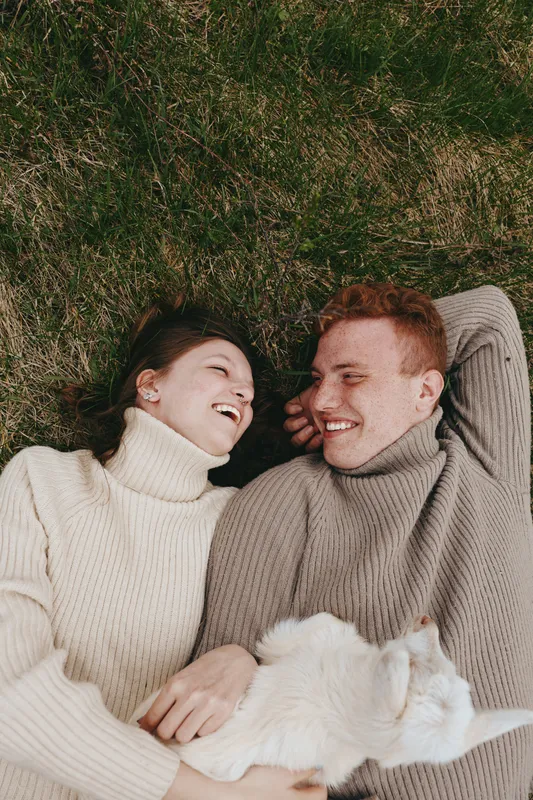 Couple in cozy sweaters lie on grass, sharing warm smiles, while a fluffy cat rests beside them, creating a tender lifestyle moment of affection, companionship and outdoor comfort.