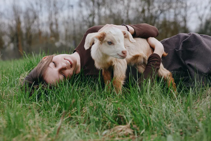 goat kid animal outdoors lifestyle nature, cheerful person lying on grass with a playful goat, relaxed mood, soft natural light, rustic countryside vibe, companionship and gentle affection, candid