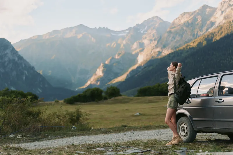 A lone young woman with a backpack stands beside a parked car in a rugged mountain valley, preparing for a hike and enjoying a moment of solitude. A feeling of independence.