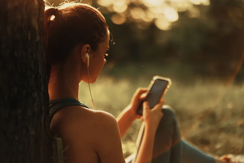 Beautiful young woman listening to music on her smartphone outdoors, surrounded by a sunset glow, showcasing a relaxed and joyful mood