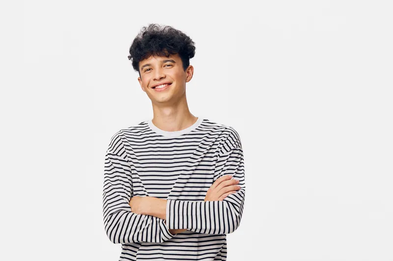 Smiling young man with curly hair wearing a striped sweater, standing confidently with crossed arms against a plain white background, casual and relaxed look.