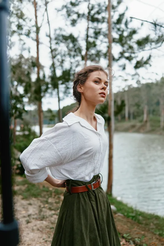 Young woman in a white blouse and green skirt standing by a river, exuding confidence and tranquility amidst a natural setting