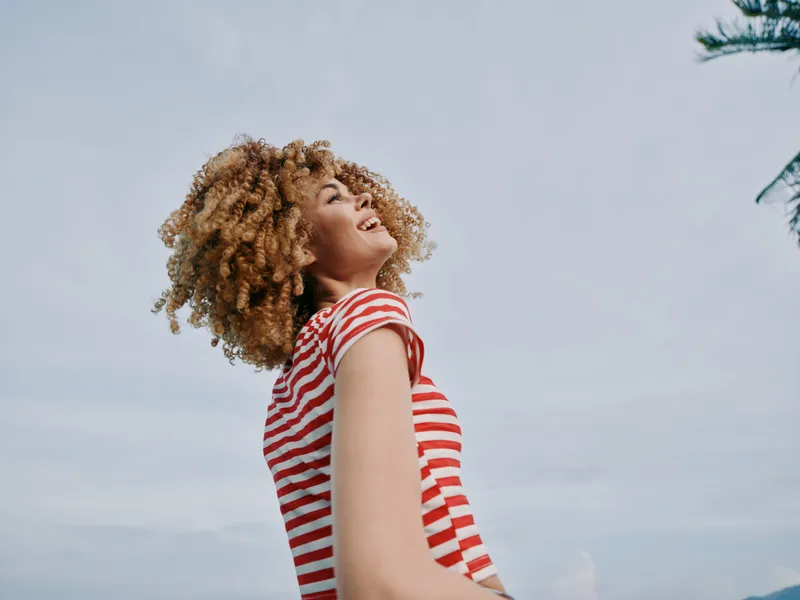 A joyful portrait of a person with curly hair wearing a red and white striped shirt, standing outdoors as wind lifts hair toward a bright blue sky.