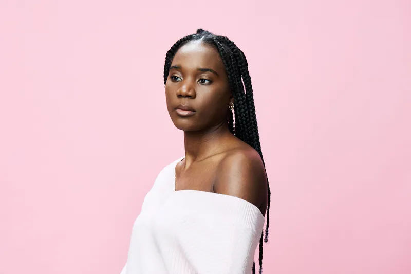 Portrait of a young Black woman with braids, wearing a soft sweater, against a pastel pink background, conveying a sense of calm and confidence