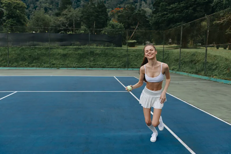 Happy young female tennis player in white sportswear running on a blue court, showcasing energy and joy in an outdoor setting