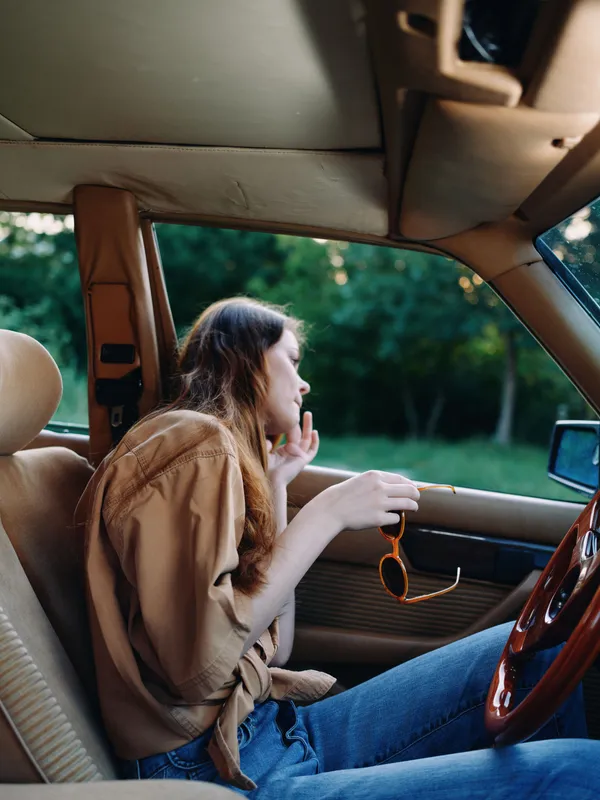 Young woman in casual beige shirt and blue jeans sitting in car, looking thoughtful with sunglasses in hand, surrounded by greenery on a sunny day.