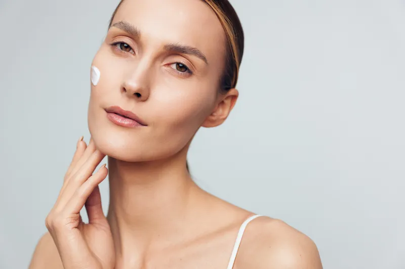 A young woman with brown hair applies facial cream to her cheek in a soft studio setting, conveying a calm skincare moment and focus on self care.