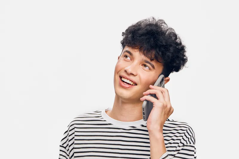 Young man talking phone smiling, casual striped shirt, curly hair, bright studio background, positive expression, communication moment, lifestyle portrait
