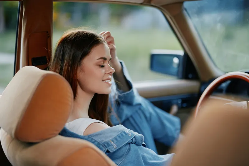 Young woman relaxing in a vintage car, wearing denim jacket, with a serene expression, showcasing a carefree summer vibe and warmth of nostalgia.