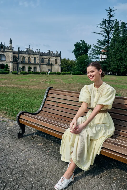A young woman sits on a wooden bench in a sunny park, wearing a pale yellow dress and white shoes, smiling with a historic castle in the background