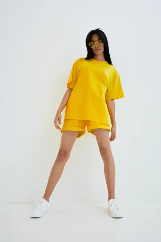 Young woman wearing bright yellow oversized t-shirt and shorts posing confidently with white sneakers and sunglasses against a plain white background in casual summer style.