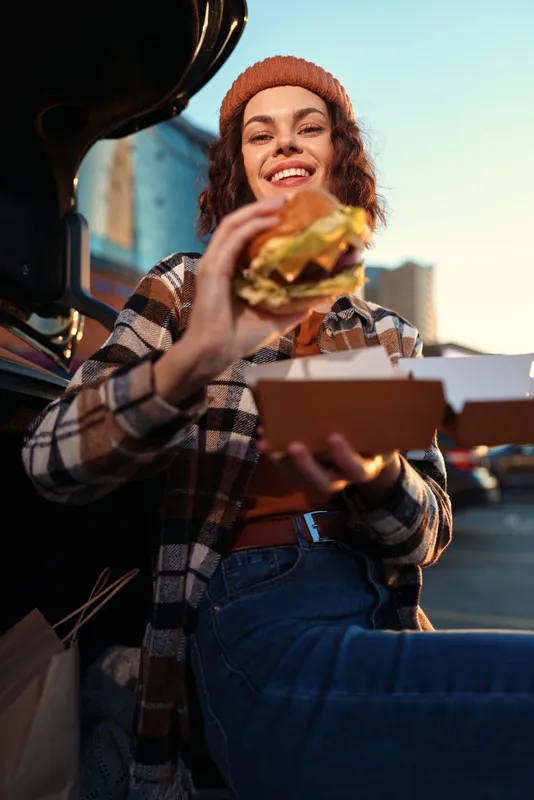 Woman with burger by car trunk, smiling outdoors at golden hour, candid lifestyle shot with authenticity and emotional storytelling, cozy urban vibe and mindful living