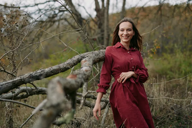 A candid portrait of a woman in a deep red dress leaning on a rustic fence in a quiet meadow, radiating authentic charm and natural presence and sincere mood.
