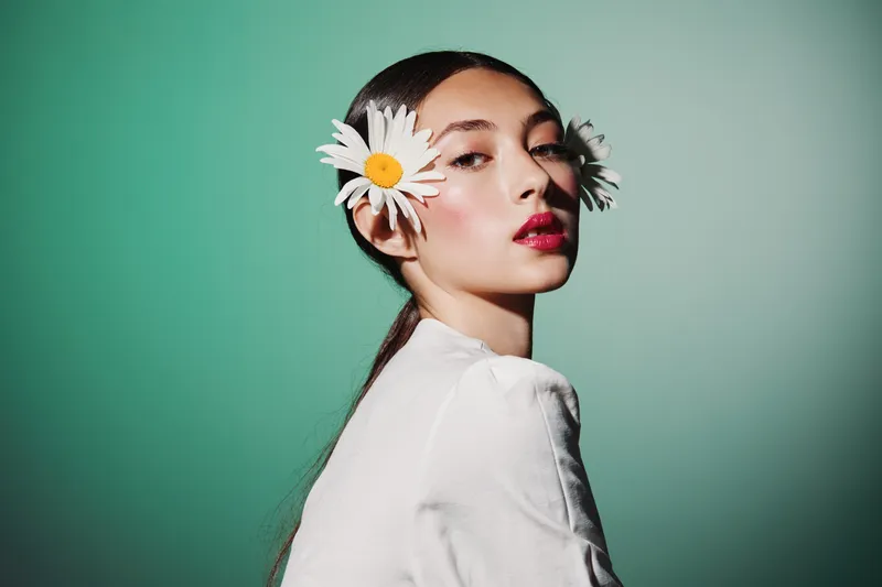 Young woman with daisy flowers in hair poses in white blouse against green background. Her skin is smooth and glossy with bright red lips. Beauty fashion concept with natural styling.