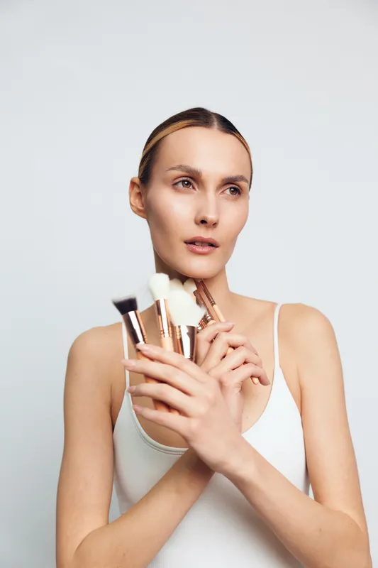 Young woman with smooth skin holding makeup brushes in both hands against a white background. Concept of beauty, cosmetics, skincare, and natural look with soft lighting.