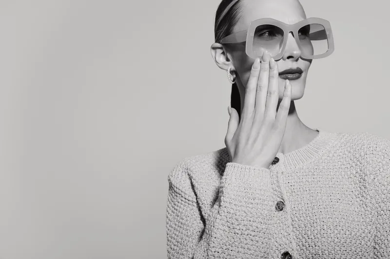 Blonde white woman wears large sunglasses and textured jacket, posing with hand near face on neutral background. Concept of fashion, style, and elegance.