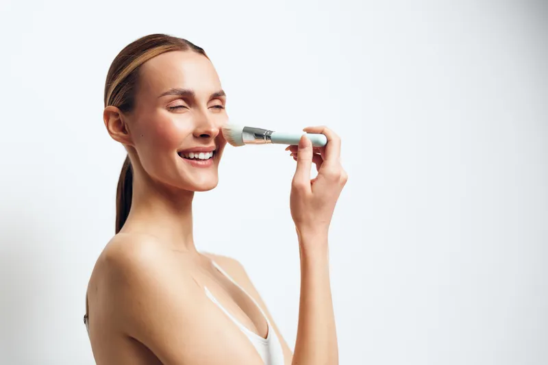 Smiling woman with tied hair applying facial serum with a dropper on clean skin. Bright studio lighting on white background. Concept of skincare, beauty, and self-care routine.