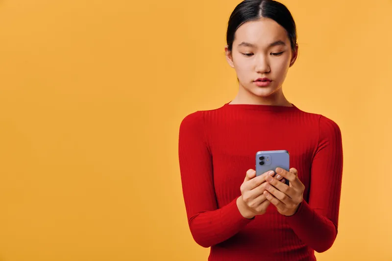 Young woman in red sweater holds smartphone and uses touch screen on orange background. Casual Asian female focused on mobile device for communication or social media.