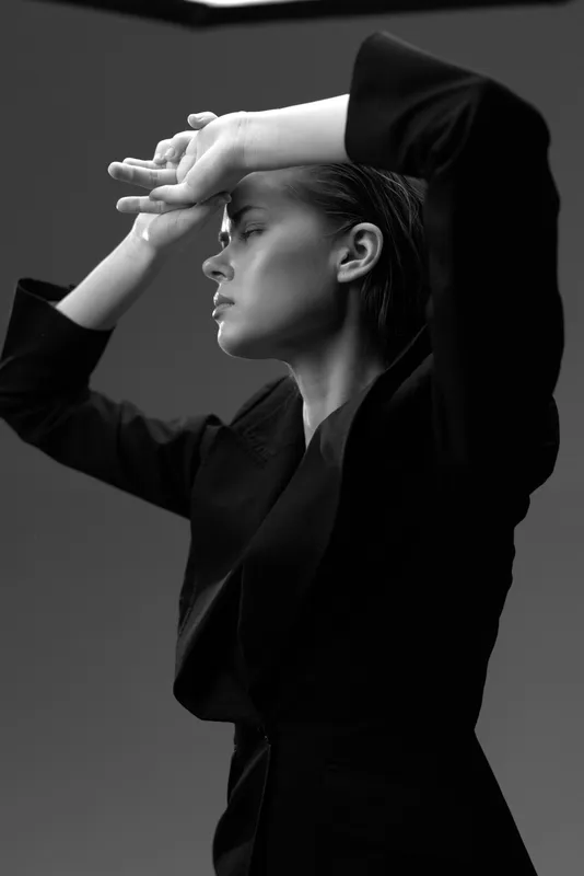 Black and white portrait of a thoughtful woman in dark clothing with hands raised, showing strong profile and serene expression.