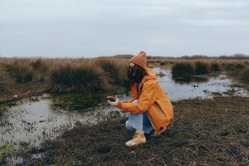 Woman in yellow raincoat with hood sits by small river in wet countryside, holding phone and enjoying nature lifestyle, cloudy sky, quiet landscape, puddle reflections, outdoors after rain