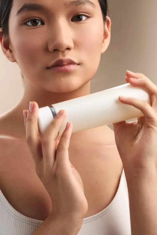 Asian woman applies skincare from a white bottle in a soft light setting, focused expression, clean beauty routine, calm and serene atmosphere.