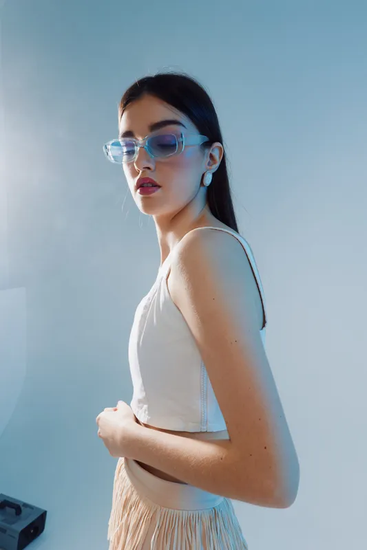 Young woman with long dark hair wearing transparent glasses and a white top poses against a blue background. Concept of beauty fashion and modern style.