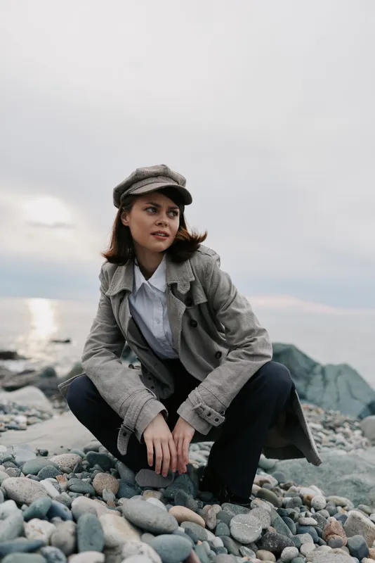 Young woman wearing a textured coat and hat crouches on rocky beach, looking away. Overcast sky and calm sea create moody atmosphere, fashion concept for autumn or winter outdoors.