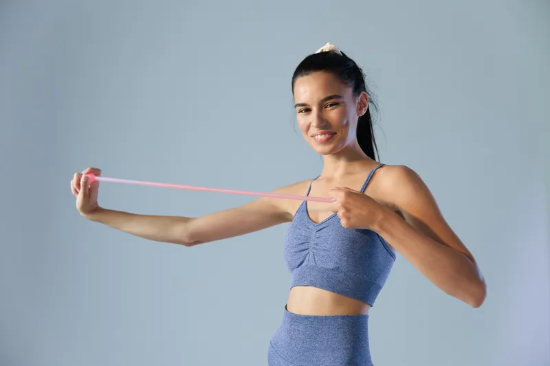 Young woman sport fitness stretching with a resistance band in blue activewear, smiling and focused against a plain light background, showcasing strength and health.