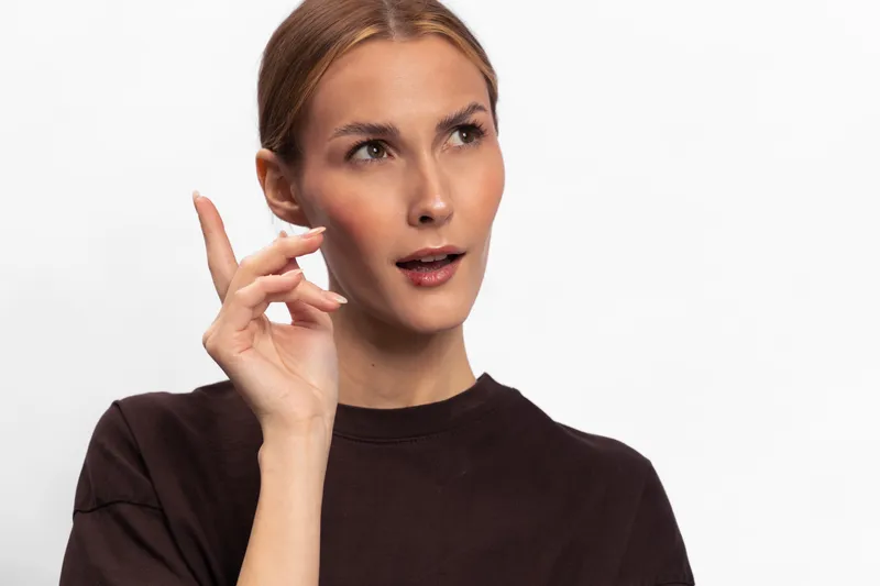 Thoughtful woman in dark shirt looks away with raised hand near face. She appears to be considering or explaining something against a plain white background. Concept of thinking and expression.