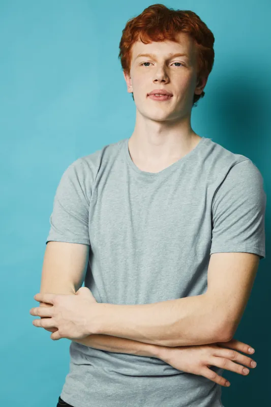 Young man with red hair wearing grey t shirt standing with arms crossed against blue background looking straight with calm expression in studio portrait.