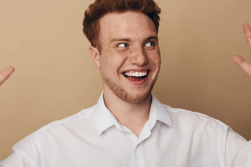 Happy young man with red hair and beard smiles broadly, wearing white shirt, raising hands on beige background. Concept of joy, excitement, and positive emotions.