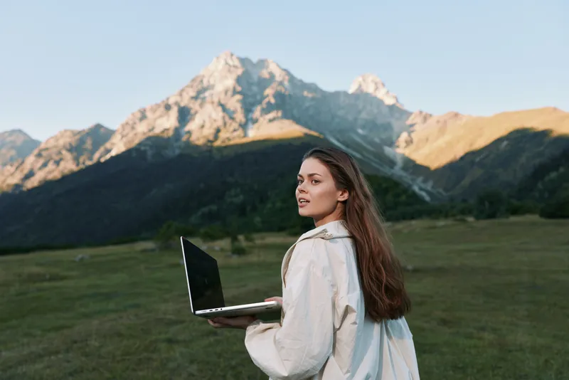 A cheerful professional woman stands outdoors with a laptop in a scenic mountain landscape. She appears focused and confident, embodying a modern remote work vibe in nature.