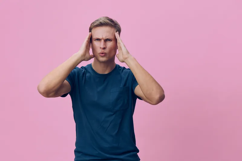 Frustrated man with short blond hair and casual blue t-shirt holding his head with hands, showing stress or confusion, studio shot on pink background, no accessories, casual clothing, emotional expres