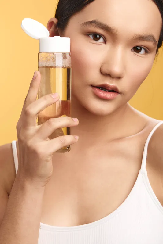 Asian woman holds a clear bottle of golden liquid in a bright studio, looking softly at the camera with a poised, confident expression, suggesting premium skincare and self care.
