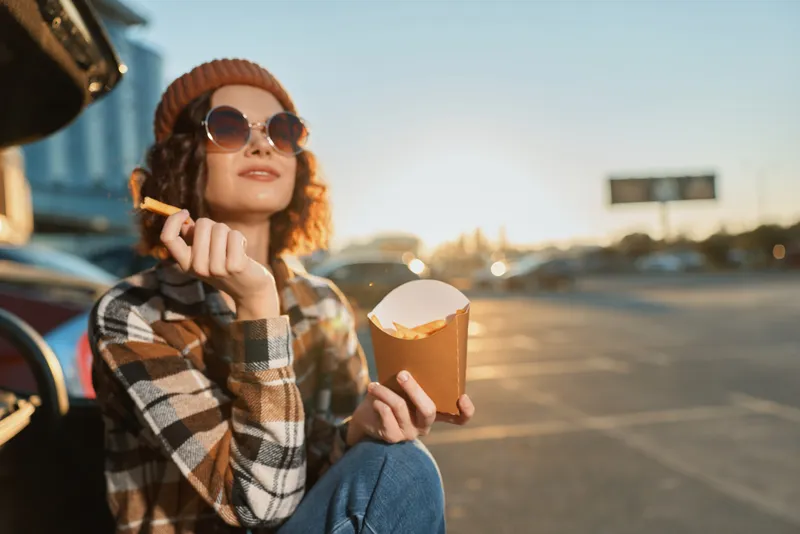 Woman eating fries by car at sunset, sunglasses and beanie, casual outdoor scene with open trunk; candid lifestyle portrait with golden hour glow, authenticity, mindful living, emotional storytelling.
