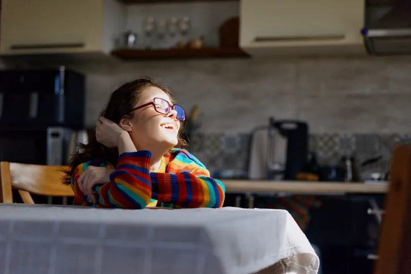 Cheerful girl indoors, smiling with glasses, striped sweater, relaxed moment at home, cozy kitchen background, soft natural light, candid lifestyle