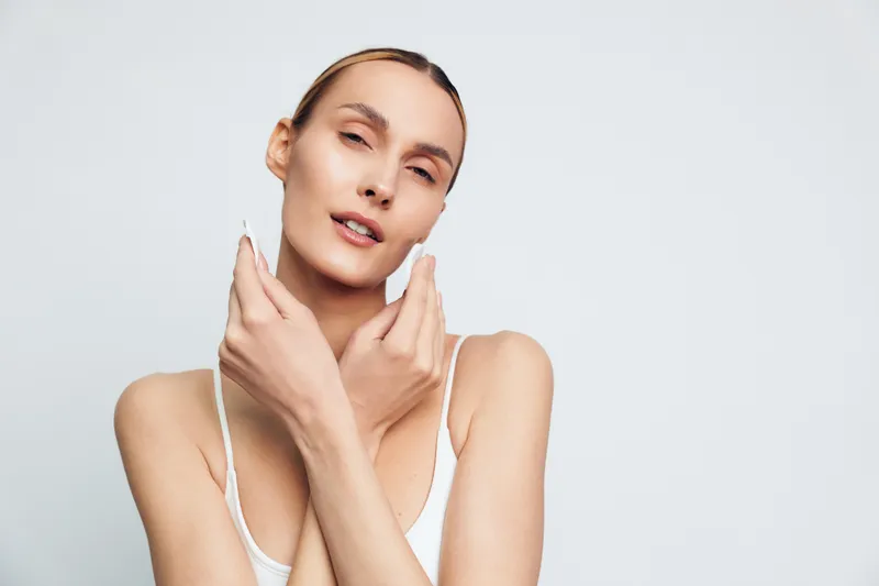 A young woman with slicked-back hair poses with her hands gently touching her neck and face while wearing a white tank top against a plain light background, expressing calm confidence.