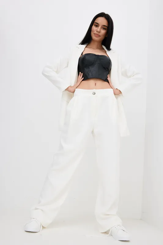Stylish woman posing confidently in white wide-leg pants and blazer with black crop top, showcasing modern fashion and casual elegance on plain light background.