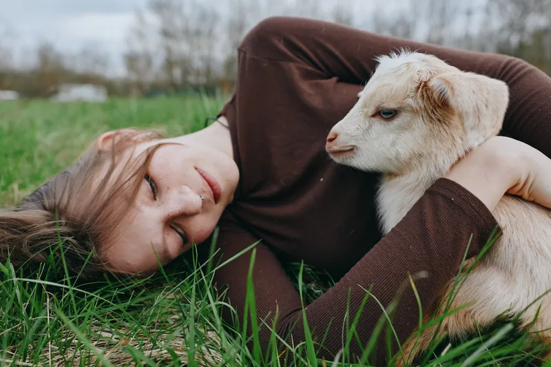 Cheerful person resting in grass with a curious goat kid, outdoor sunlight, relaxed posture, intimate countryside moment, natural colors, gentle breeze, lifestyle storytelling