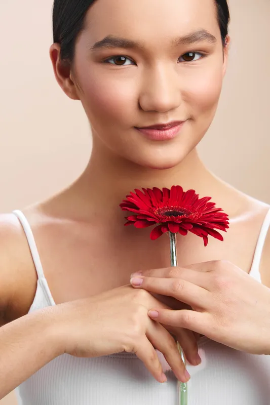 asian woman smiles softly while holding a red flower close to her chest in a clean studio setting, conveying calm beauty, gentle confidence, and graceful presence.