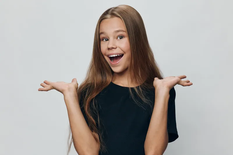Girl with long hair smiling and showing surprised expression with raised hands on gray background. Child with casual black t shirt posing happy and cheerful in studio lighting.