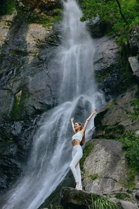 Young woman in white activewear stands joyfully near a tall waterfall surrounded by lush greenery and rocky cliffs, embracing nature’s beauty and adventure.