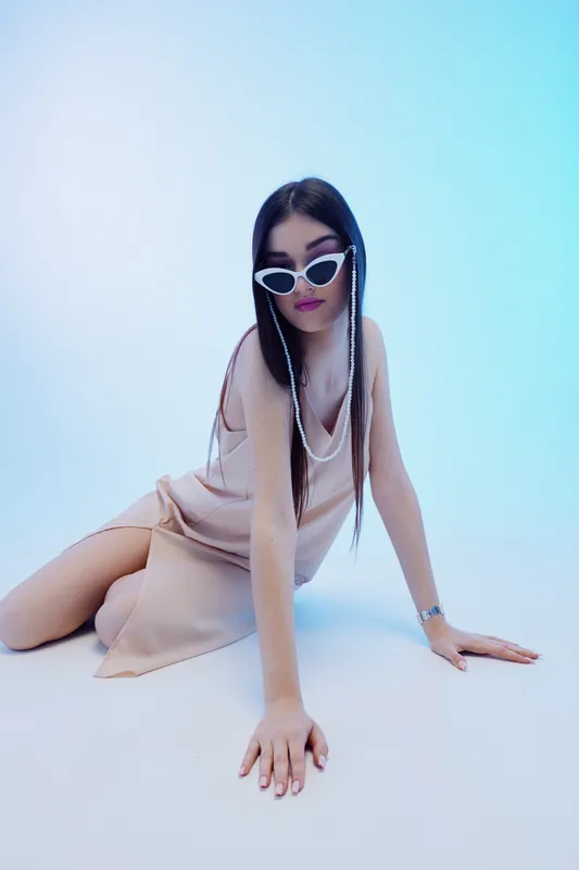 Young woman in stylish beige dress and white sunglasses poses on floor in blue studio light. Concept of woman beauty fashion and modern elegance.