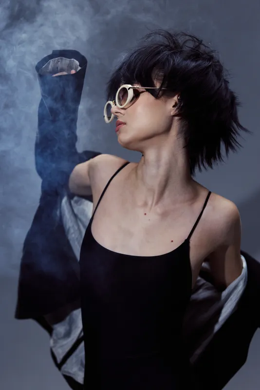stylish woman in sunglasses with black outfit posing with smoke against dark background