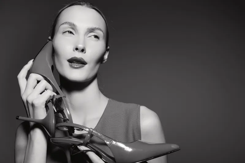 Portrait of a young white woman with pale skin holding red high heel shoes near face, isolated on dark background, fashion and beauty concept in black and white.